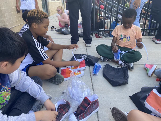 Children sitting on a concrete floor outdoors, trying on colorful sneakers and organizing shoe bags around them.
