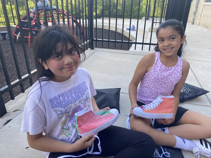 Two children sitting on a concrete surface near a playground, each holding a pink and orange high-top sneaker.
