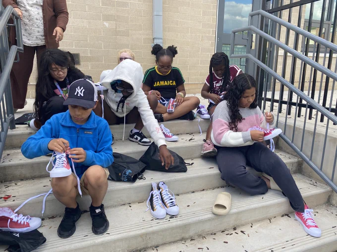 Group of children sitting on outdoor steps, focused on tying white sneakers with red and blue accents during a casual activity.
