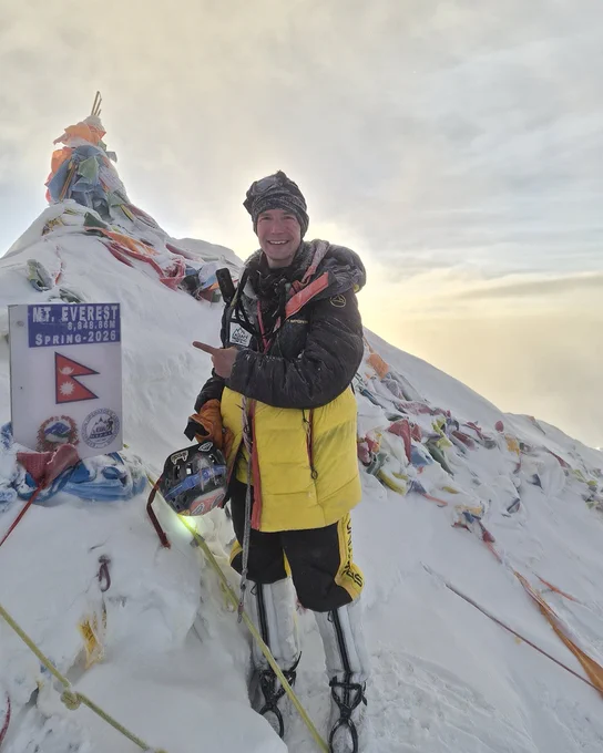 Tyler Andrews stands atop Mount Everest wearing a black and yellow winter jacket and pants, as he points to a sing that reads "Mt. Everest 8,848.86M Spring 2026" 