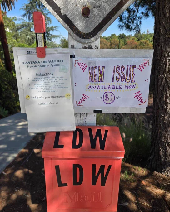 Outdoor neighborhood newspaper stand labeled “LDW” painted bright orange, positioned beneath a street sign. Above the box, two posted sheets include instructions for a $1 honor‑system newspaper purchase and a colorful sign reading “NEW ISSUE AVAILABLE NOW $1.” The stand sits beside a sidewalk with trees and greenery in the background under sunny daylight. 