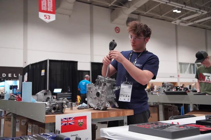 Student using a handheld tool works on a metal engine component on a workbench at a technical competition.
