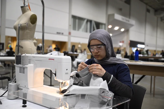 Student wearing safety glasses operates a sewing machine, carefully stitching gray fabric in a workshop setting.