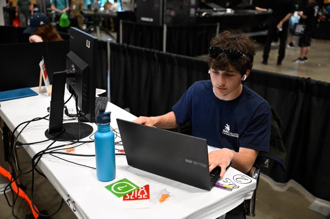 Student sits at a table using a laptop connected to a vertical monitor, with cables, a water bottle, and tools nearby in a workspace environment.