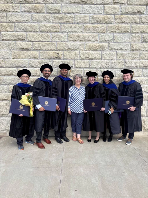 Kim smiling amid graduating K-State Physics PhD students in their caps and gowns after the spring commencement ceremony.