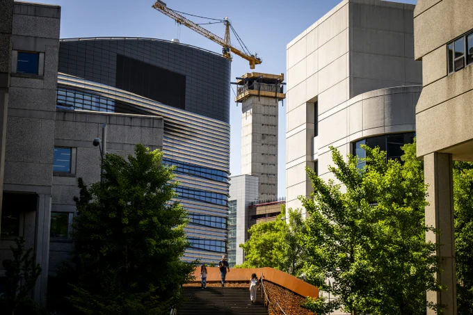 Modern buildings and a yellow construction crane against a clear sky. Below, three people walk down stairs with an orange railing, surrounded by green trees. A tall concrete structure is under construction in the background.