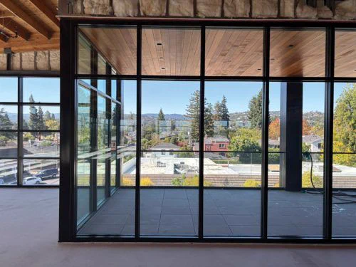 View through large glass walls of a modern building, showcasing a scenic landscape with homes, trees, and distant hills under a clear sky.