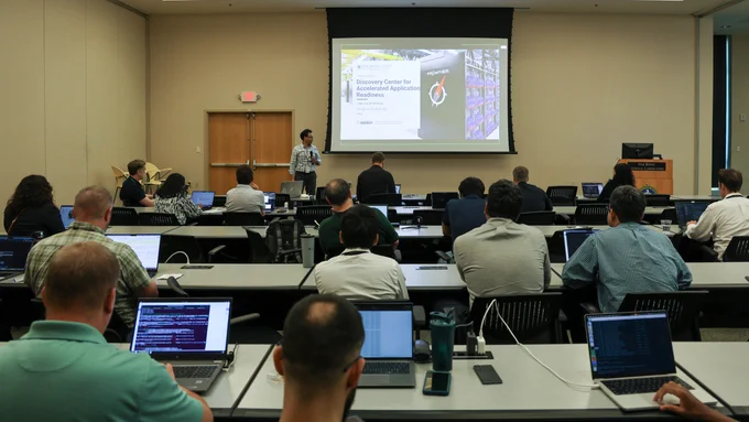 A presenter stands before a projected slide titled “Discovery Center for Accelerated Application Readiness” while addressing workshop participants seated throughout a conference room with laptops open.