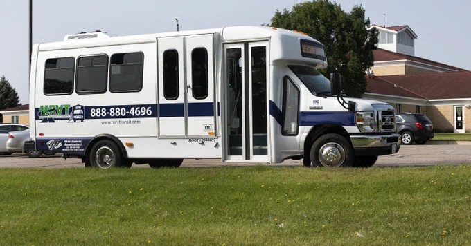 A white transportation bus is parked on a grassy area near a building with a sloped roof. The bus has Minnesota River Valley Transit branding and a contact phone number displayed on its side.