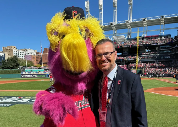 A person standing on a baseball field is smiling with the Cleveland Guardians mascot, Slider. The mascot has purple fur and wears a team baseball cap. The background features Cleveland's baseball stadium, Progressive Field, with spectators and banners.