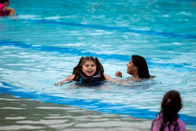 Two people are swimming in a pool, with one wearing a life jacket. Another person is near the edge of the pool observing them.