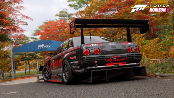 A sleek black and red racing car is parked on a scenic road with autumn foliage. The car features prominent logos and branding like "GT Xtreme" and "PRP." In the background, a blue archway displays "Hokubu Exclude." The image is from "Forza Horizon 6.