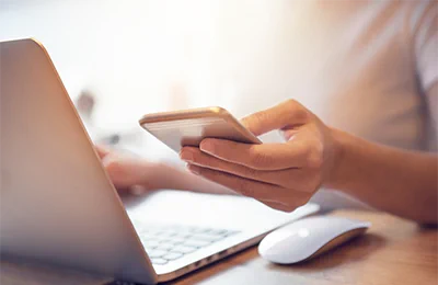 Person holding a smartphone in one hand while working on a laptop with a wireless mouse nearby on a wooden desk.