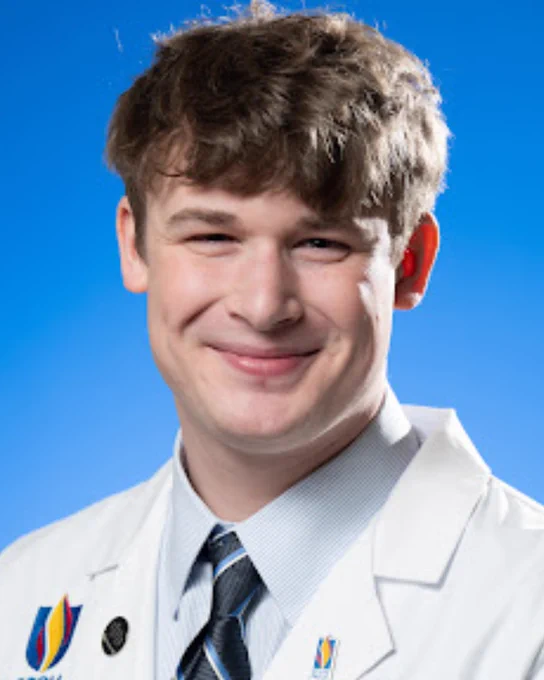 A person smiling at the camera in front of a blue background. They are wearing a white coat business professional clothing. 