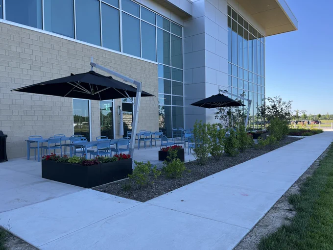 A modern building with large glass windows and a light beige exterior. An outdoor patio is furnished with blue chairs and black umbrellas, surrounded by planters with green shrubs and red flowers. A concrete walkway leads alongside a grassy area, under a clear blue sky.