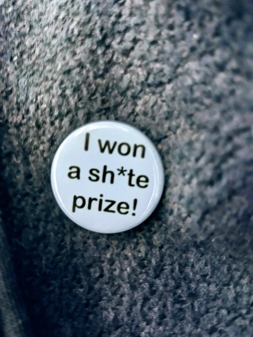A round, white, metal pin badge with black writing on it, which says, "I won a shite prize!"