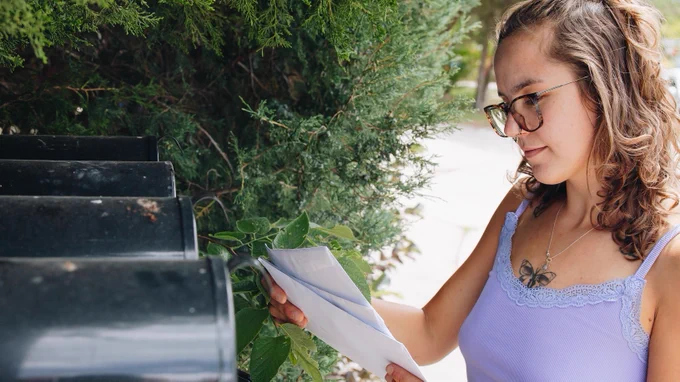 A woman holds multiple letters in front of a row of mailboxes.