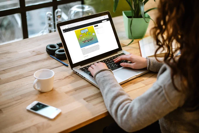 Female working on laptop with her hands over the keyboard. She is looking at the In Dialogue/En Diálogo website. Cell phone, coffee mug, headphones, pen, book, and plant are also on top of the desk.
