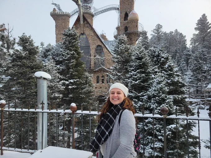 Lucia Rudela in front of snow-covered pine trees surrounding a whimsical castle with turrets under a gray winter sky