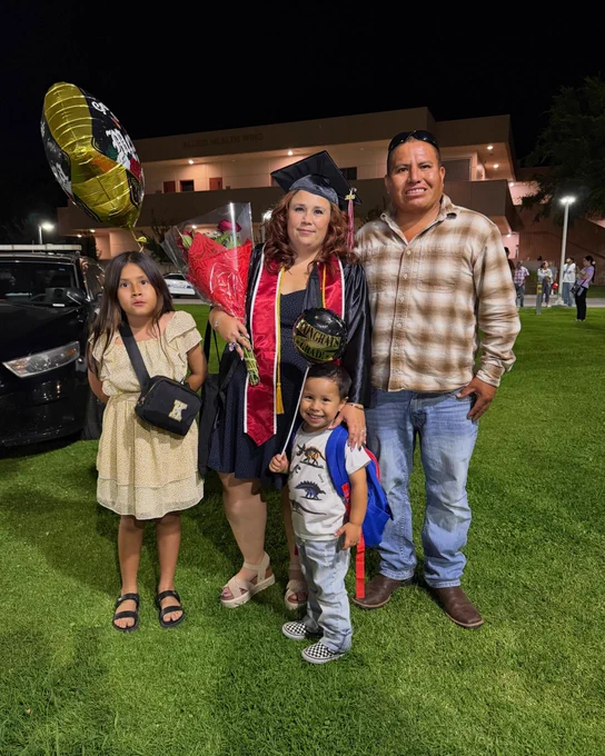 Tiana Hinojosa in graduation cap and gown holding flowers and balloon, standing with family on grass at night outside a building