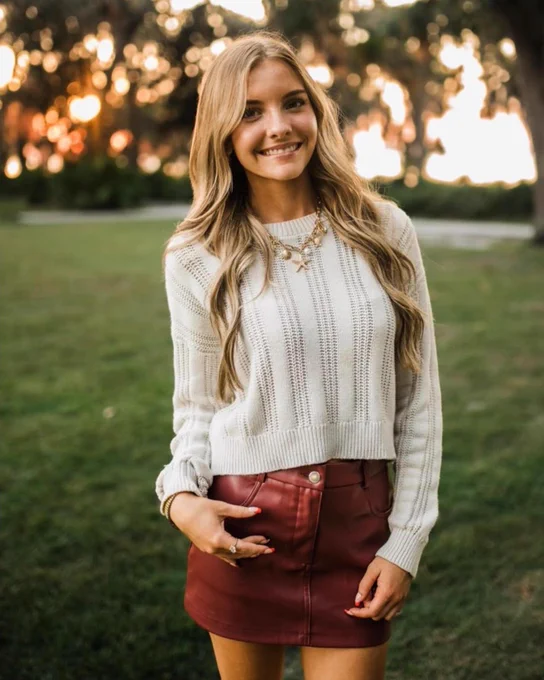 Addyson Davis wearing a white knit sweater and a red leather skirt standing outdoors with trees and sunset in the background