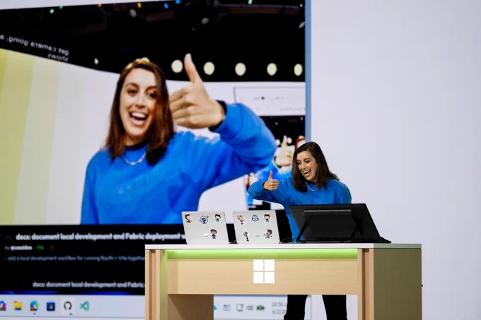 Una persona con un suéter azul da un pulgar hacia arriba desde un escenario. En el fondo, hay una pantalla grande que muestra texto sobre 'desarrollo de documentos locales y despliegue de Fabric.' Un logo de Windows es visible en un escritorio.