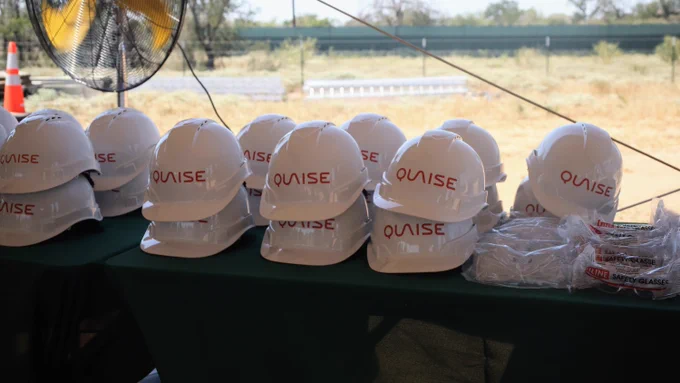 A row of white hard hats labeled "Quaise" is displayed on a green tablecloth outdoors. Nearby, clear plastic packaging and a safety cone are visible in the background.
