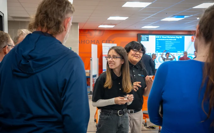 Two FAME students standing before a group of audience members after their final presentations. One has long, brown hair and the other has short, dark hair. They both wear their black FAME polos.