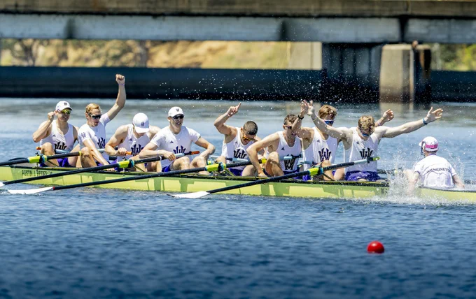 A men's rowing team in a yellow boat on blue water, celebrating with raised arms and fists. They wear white tops with a purple 'W' logo. Text on boat: \"Austin J. Regier\". A coxswain (WASHINGTON) is at the back. Water is splashing. A red buoy is in front.