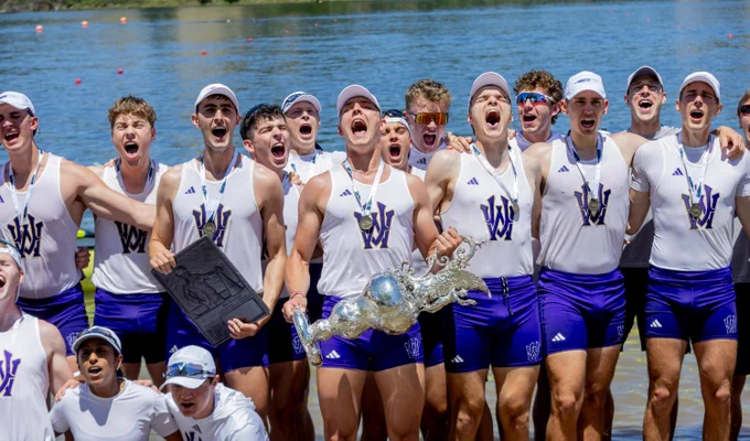 A large group of excited young male rowers, wearing white tank tops with a purple 'W' logo, purple shorts, and medals, cheer loudly. They hold a large, ornate silver trophy cup and a black embossed plaque, standing by blue water.