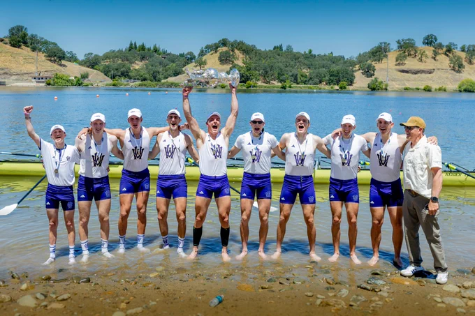 Nine young male rowers and an older man stand jubilantly in shallow lake water. The rowers wear white shirts, purple shorts with a 'W' logo, and medals. One proudly hoists a large silver trophy. A yellow rowing shell is behind them, with green hills and a blue sky in the background.