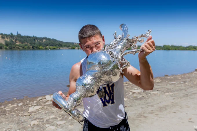 A young man, wearing a white tank top with a blue and gold 'W' logo, holds a large, ornate silver trophy to his face. He is on a rocky shore next to a calm lake, with green hills and a clear blue sky in the background.