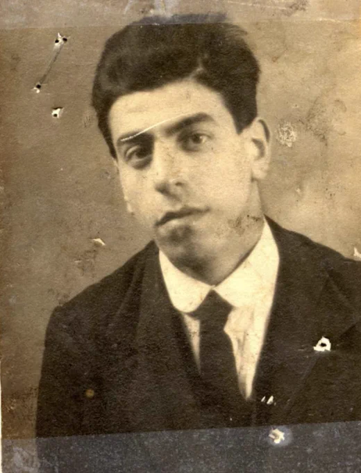 Vintage sepia-toned portrait of a man wearing a dark suit and tie. The photograph shows some signs of wear, including small tears and marks. The person has a serious expression and styled hair. The background is a plain, indistinct surface.
