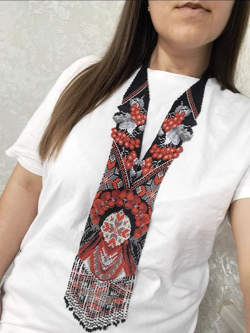 A close-up photo of a woman wearing a traditional Ukrainian beaded necklace called a gerdan over a plain white t-shirt. The gerdan is hand-woven with black, red, white, and grey beads. It features intricate patterns of red viburnum (kalyna) berries with grey leaves on the sides, leading down to a large central geometric ornament resembling a stylized girl figure (Berehynia symbol). The necklace ends with long, detailed beaded fringes at the bottom.