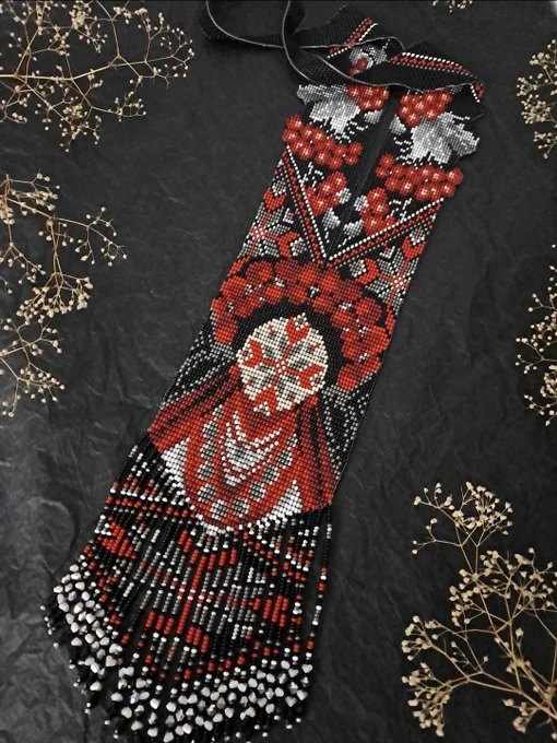A top-down view of a traditional Ukrainian beaded gerdan necklace laid flat on a textured black background. The handmade necklace features black, red, white, and grey beadwork with patterns of red viburnum berries and grey leaves. The central part displays a large geometric motif representing a stylized girl figure (Berehynia). The bottom of the gerdan is finished with long beaded fringes. Small, delicate sprigs of dried white flowers are elegantly arranged around the edges of the frame.