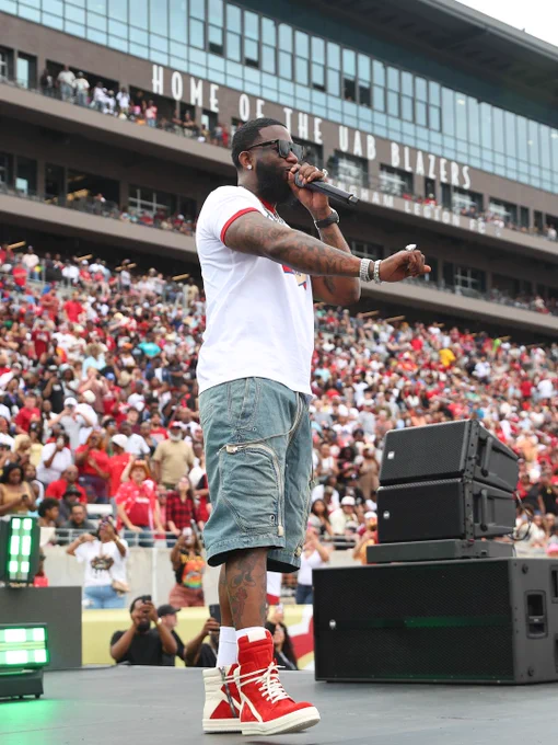 A performer on stage at a crowded sports stadium, creating an energetic atmosphere. UAB Blazers home sign visible in the background.