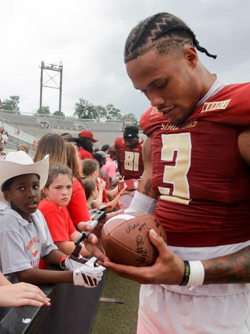 A football player in a red jersey signs a ball for fans, including children in the audience, highlighting a fan interaction moment after a game.