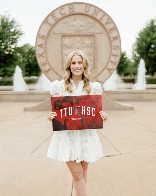Female pharmacy student holds up a sign that reads, “TTUHSC Pharmacy,” while standing outside in front of the TTU university seal.