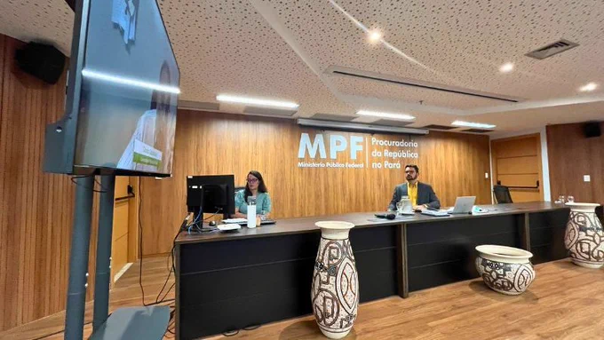 Foto de auditório com duas pessoas em bancada sob painel do MPF Pará, vasos marajoaras e TV em primeiro plano.