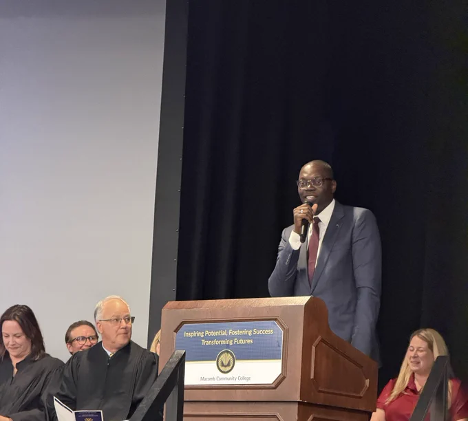 The lieutenant governor stands at a wooden podium speaking into a microphone. He wears a grey suit, white shirt, red tie, and glasses. The podium features a sign that reads "Inspiring Potential, Fostering Success, Transforming Futures" above the Macomb Community College logo. Seated on stage behind him are several individuals, including a man in judicial robes and a woman in a red dress. In the foreground, the back of an audience member's head is visible.