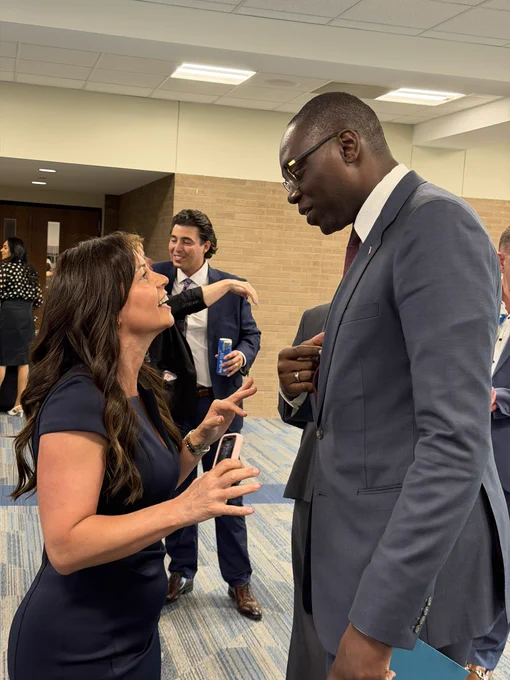 A side-view medium shot of Judge Tanya Grillo looking up and speaking with the Lieutenant Governor, wearing a blue suit, glasses, and a red tie. Judge Grillo wears a dark navy blue dress and gestures with her hands while holding a pink phone. In the background, another man in a suit smiles and holds a blue can, while other individuals mingle in a large room with brick walls and patterned carpeting.