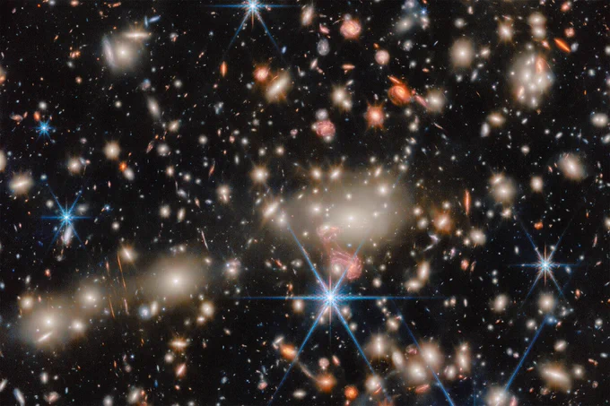 A breathtaking image of a galaxy cluster. Numerous galaxies of varying sizes and colors are scattered across the dark expanse of space, with some displaying spiral shapes and others appearing as glowing, elliptical forms. Bright stars with diffraction spikes are also visible throughout the scene.