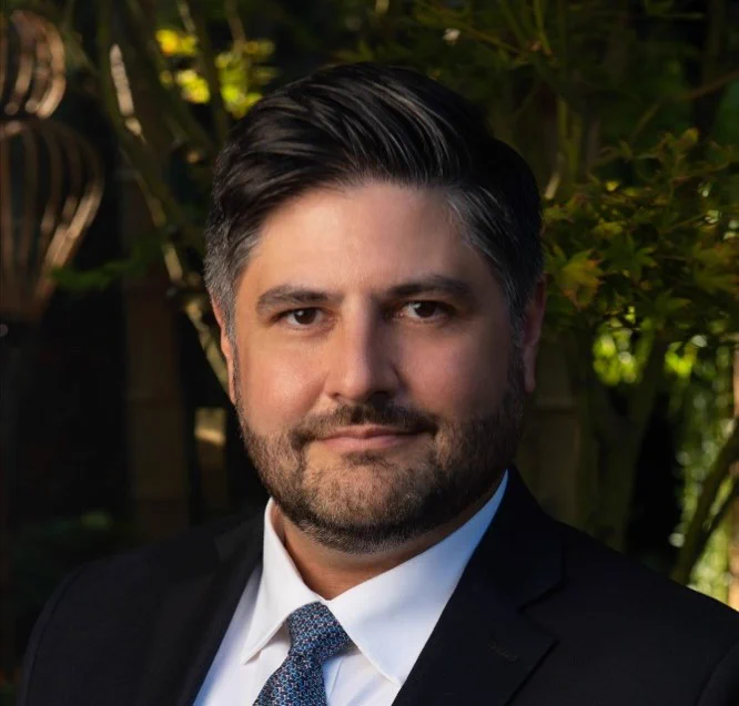 Professional headshot of a man wearing a dark suit, white dress shirt, and blue patterned tie. He is facing the camera with a slight smile. The portrait is taken outdoors with softly blurred greenery and warm natural lighting in the background.