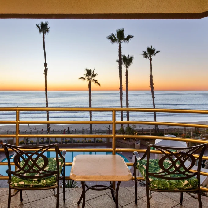 Balcony with two chairs and a table overlooking the ocean at sunset. Palm trees line the beach, and a pool is visible below.