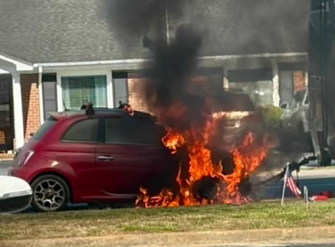 A red car is on fire near a residential house, with flames and dark smoke rising. An American flag is visible nearby on the grass.