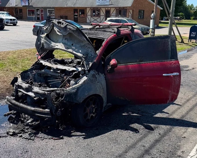 A badly burnt red car with extensive fire damage is parked on the roadside. The front end is severely charred and parts are melted. A small strip mall is visible in the background under a clear sky.