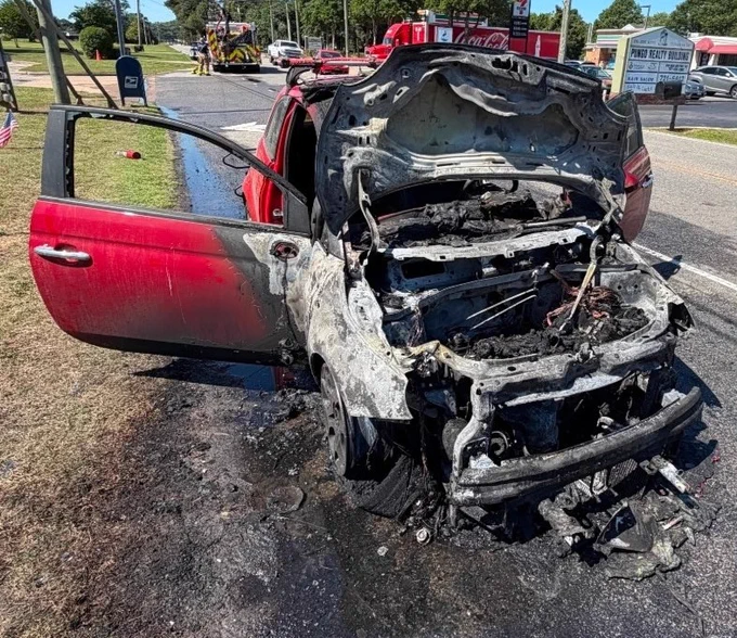 A severely burned red car is parked on the side of a road with its hood open and extensive fire damage visible. Emergency vehicles and personnel are in the background. The asphalt surrounding the vehicle is scorched.