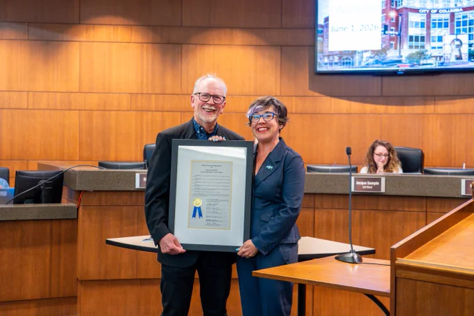 Two individuals standing in a wood-paneled council chamber holding a framed certificate with a blue ribbon.
