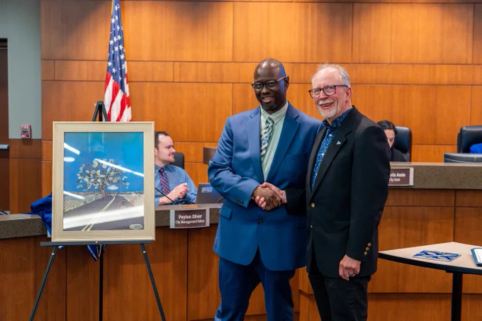 Two men shaking hands in a courtroom with an American flag and framed photo on an easel nearby.
