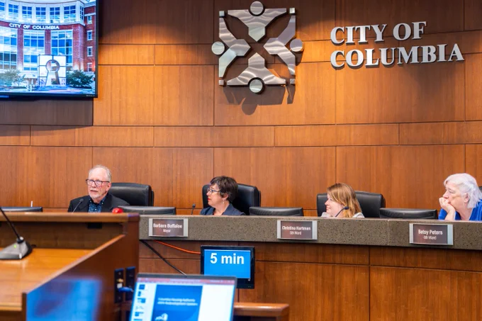 City of Columbia council members seated behind a wood-paneled dais during a meeting, with the city emblem and name displayed on the wall.